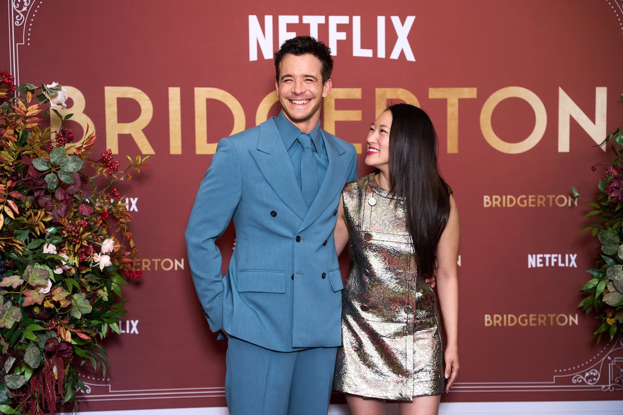A man in a blue suit and woman in a gold dress embrace and smile in front of a sign that says Netflix, Bridgerton.