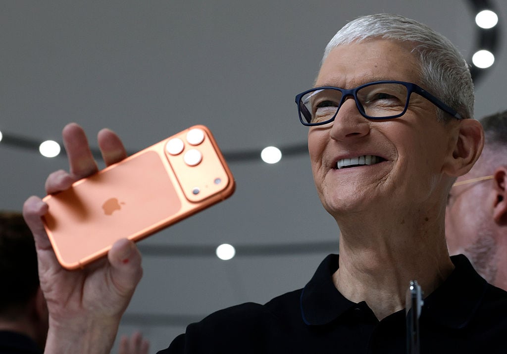A man with short, grey hair and glasses holds a copper-coloured smartphone.