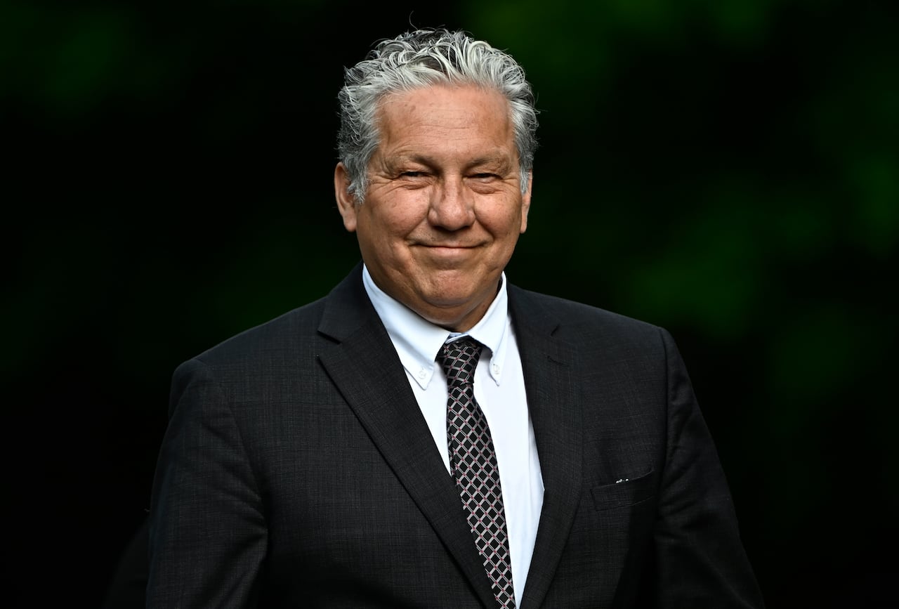 A man in a dark suit and tie smiles while standing against a black backdrop