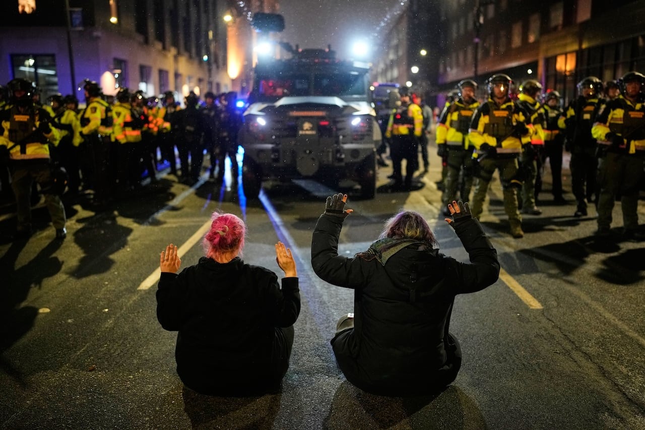 A big truck moves towards two protesters sitting on the street with their hands up, surrounded by people in helmets and uniforms