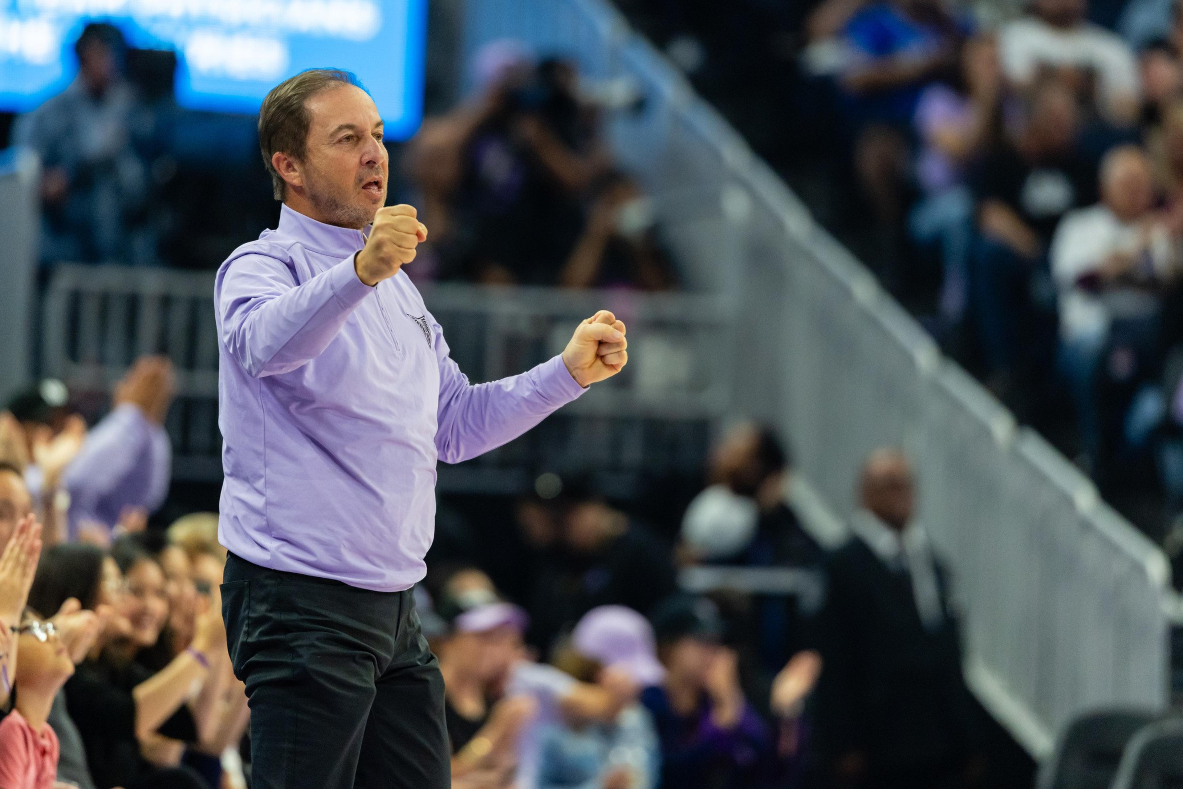 A man in a light purple shirt and black pants clenches his fists and appears to be shouting, standing in front of a cheering crowd.
