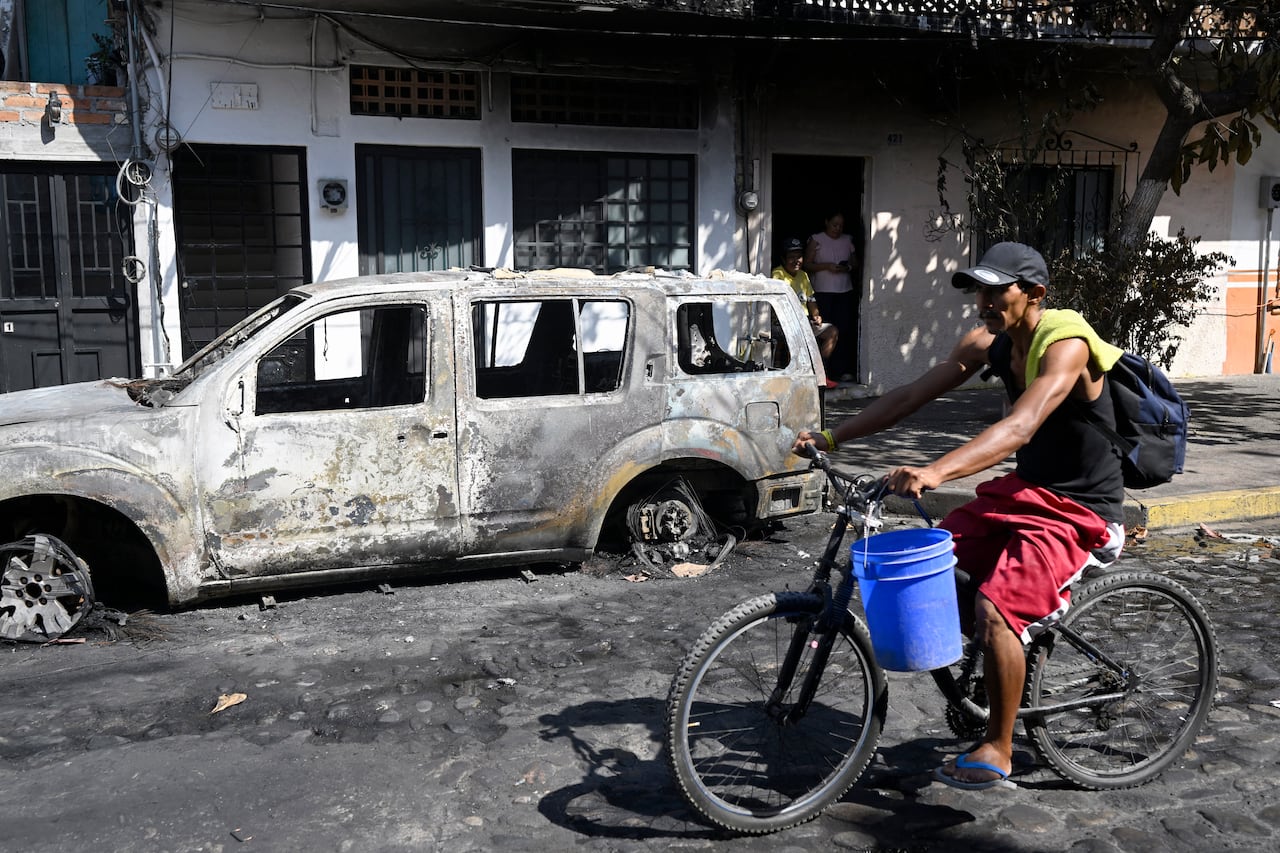 A man riding a bicycle past a burned-out vehicle