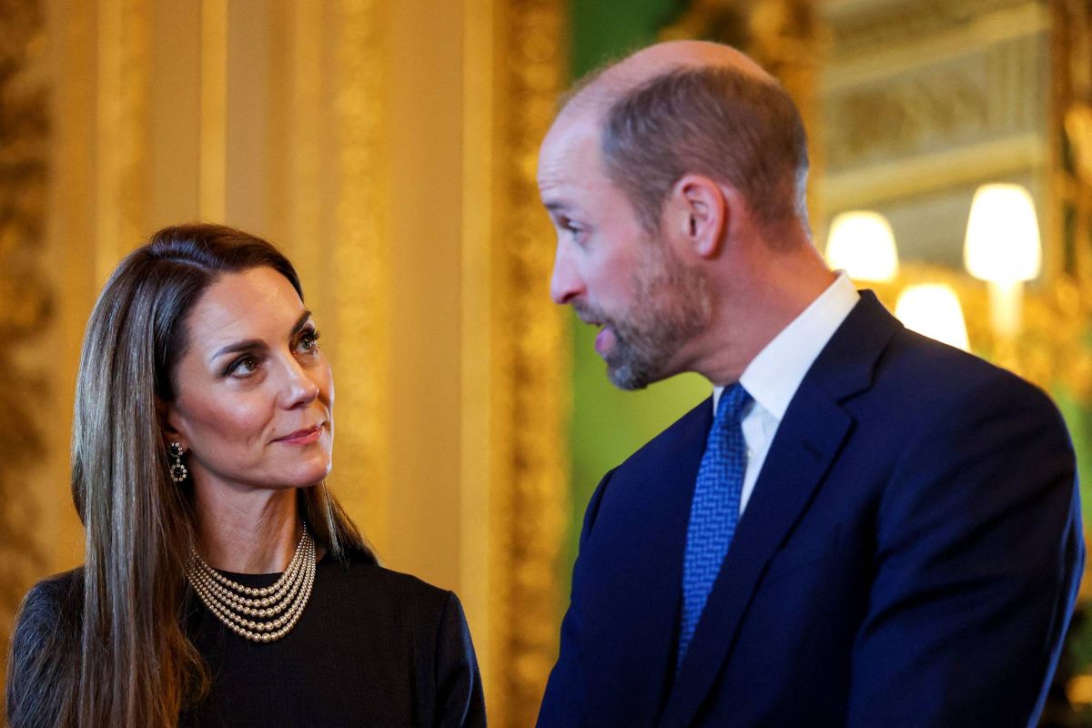 An image depicting a formal event where a man and a woman, both dressed in business attire, are engaged in a conversation. The woman, wearing a necklace, is looking towards the man who is looking towards something on her left. The background shows a decorated wall with lights.