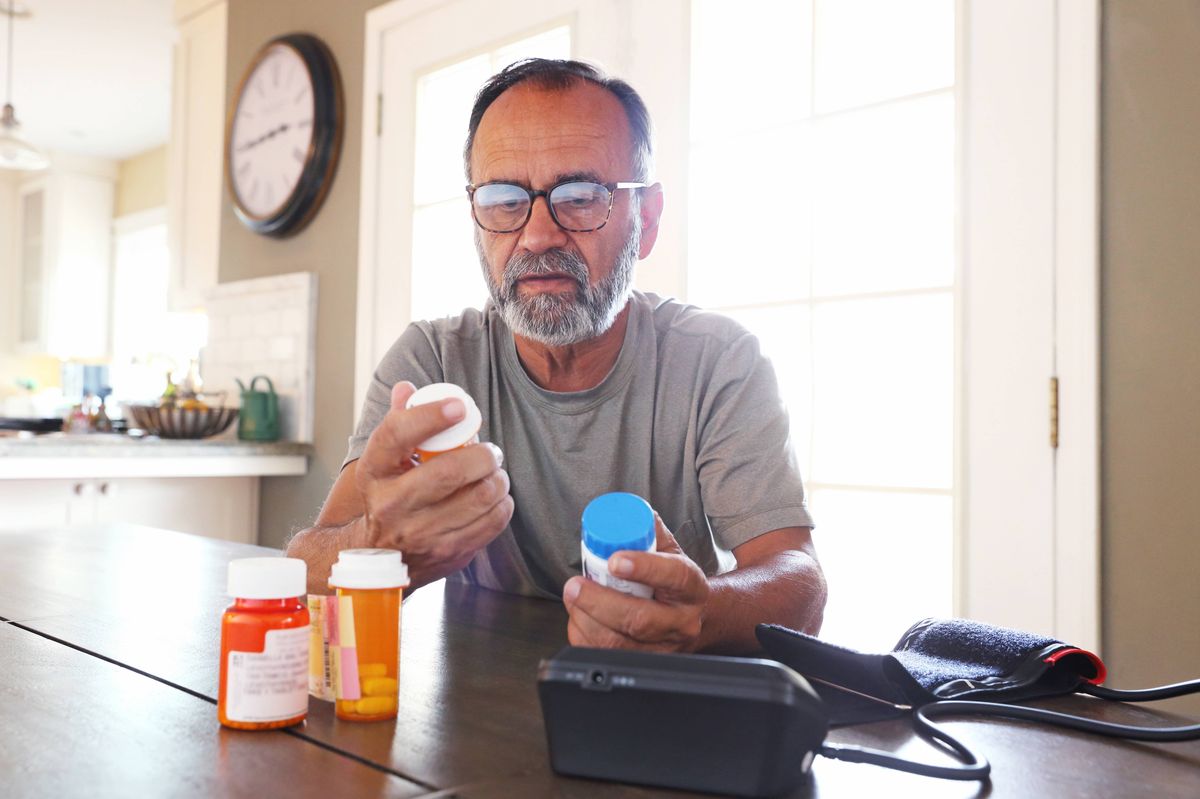 A Latino senior citizen sits at his dining room table and sorts through various prescription medications as sunlight filters in through the window behind him bathing the room with a soft glow of light. A blood pressure machine rests on the table in front of him.