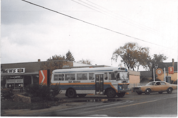 An old-looking bus is stopped at an intersection. A 1970s-era sedan turns towards the bus.
