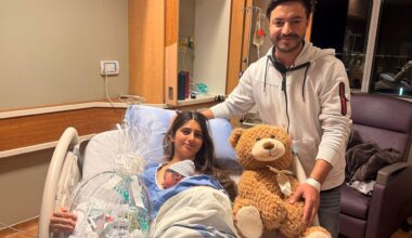 In a hospital room two parents, one holding a newborn in a hospital bed on the left and one standing to the right of the hospital bed holding a teddy bear toy
