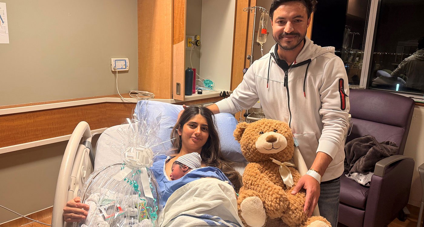 In a hospital room two parents, one holding a newborn in a hospital bed on the left and one standing to the right of the hospital bed holding a teddy bear toy