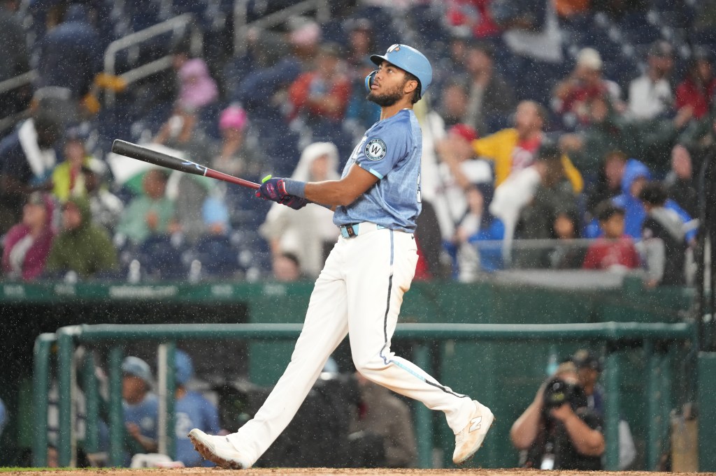 A baseball player in a light blue uniform and batting helmet looks up at a hit ball as rain falls.