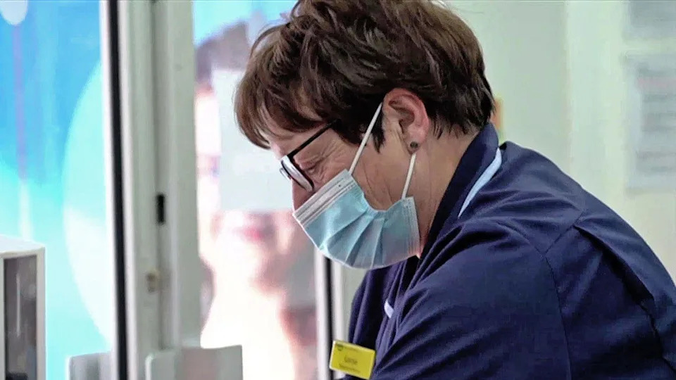 A female nurse with brown short hair wearing blue uniform and a blue disposable mask looking down. She is wearing glasses