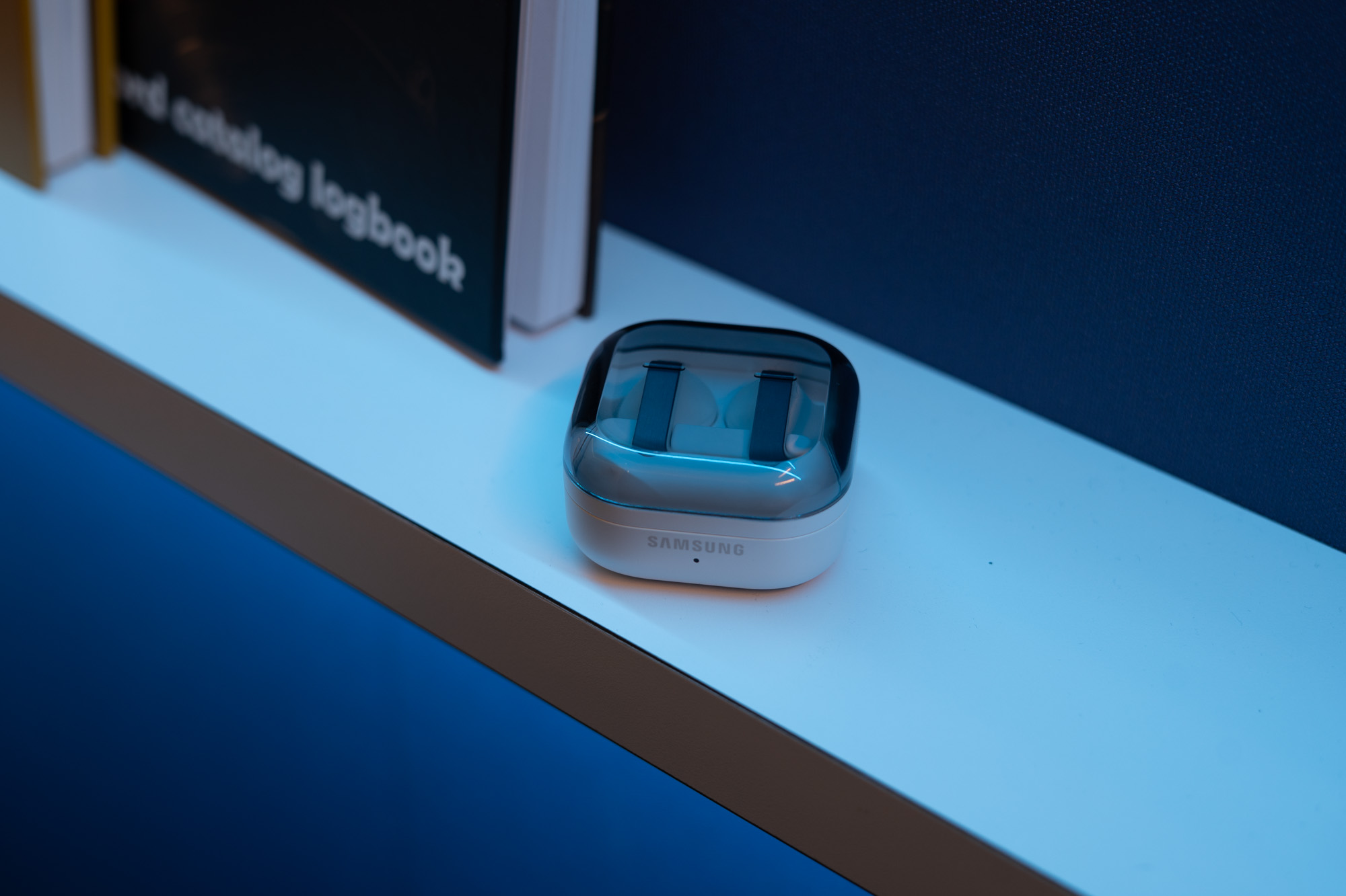Samsung Galaxy Buds 4 Pro in charging case with transparent lid on a white shelf next to a book.