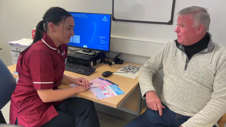 Stephanie Todd (left) is pictured in a clinical environment speaking to a patient. She is smiling and looking toward him. She has long brown hair tied up and is dressed in maroon medical scrubs. She is pictured in a clinical environment. Opposite her is Patrick Malone. He has short grey hair and is dressed in a black t-shirt with a light grey zip up jacket on top. He's sitting on a grey sofa. 