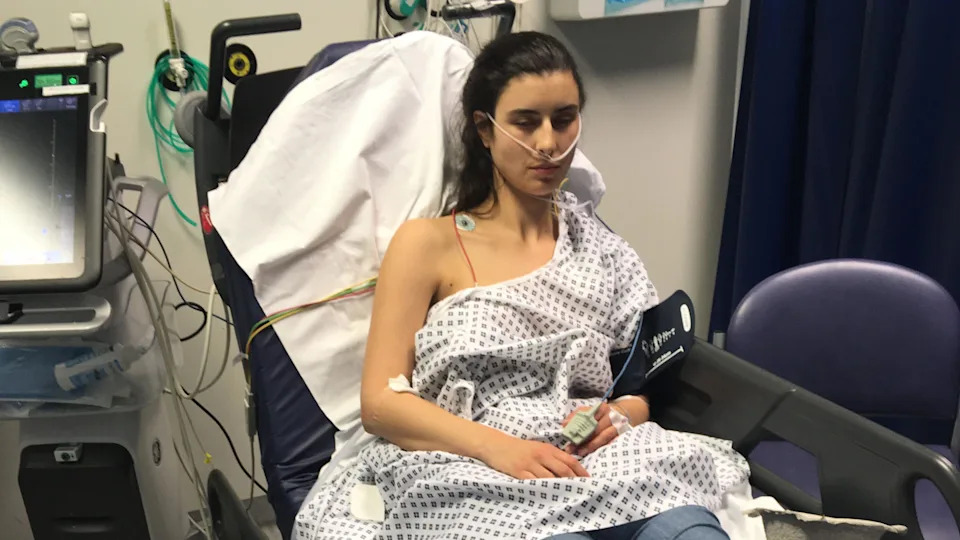 Harriet Davidson wearing a hospital gown in a hospital bed. She has her right upper chest and arm exposed and is hooked up to various pieces of medical equipment.