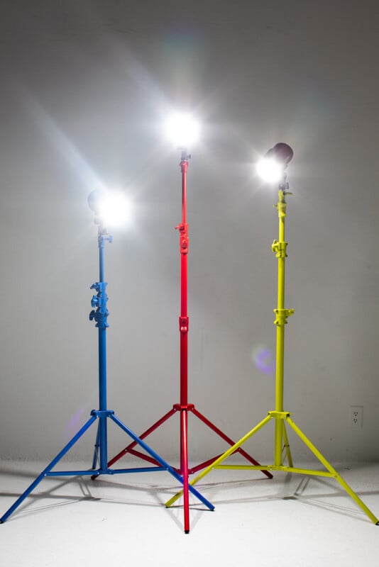 Three photography light stands in blue, red, and yellow are set up indoors. Each stand holds a bright, lit spotlight, casting strong white light against a plain, light-colored wall and floor.