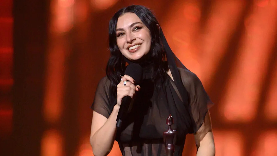 Charli XCX smiling while accepting one of her five Brit awards on stage last year. She is wearing a sheer black dress.