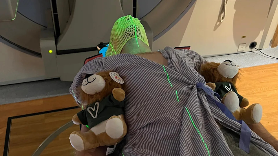 Anthony Perriam is lying in a hospital bad with two teddy bears by his side. He is wearing a mask as he is about to be scanned for cancer.