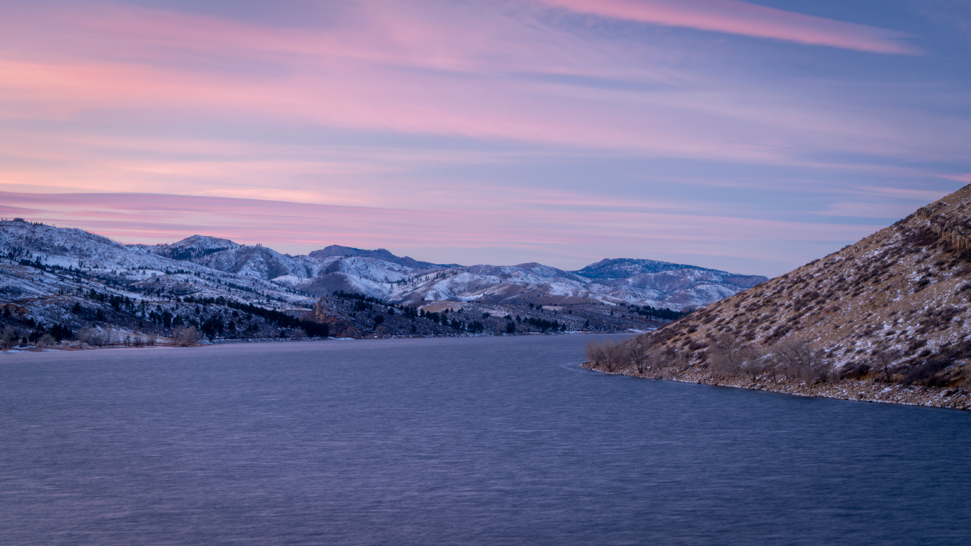 A pink sunset overlooks a large lake with sloping rocky bands on each side of the dark blue water