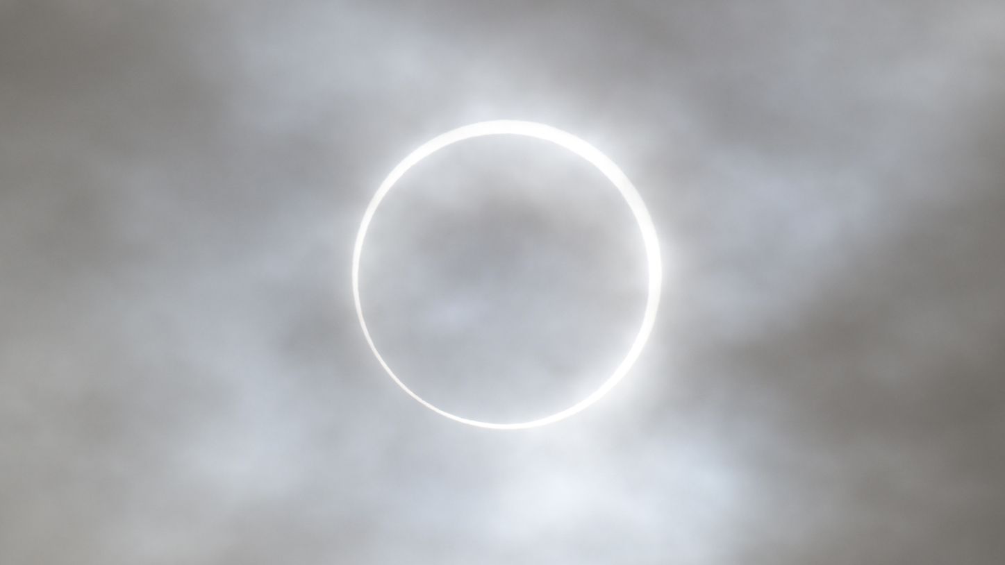 The edge of the sun is visible around the silhouette of the moon during an eclipse, forming a burning ring behind a veil of thin clouds.