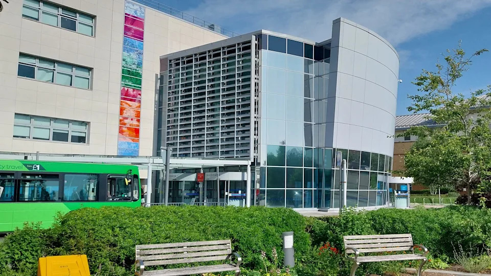 A hospital building with a green hedge and benches in the foreground. A green bus is also visible. There is blue sky and some cloud above.