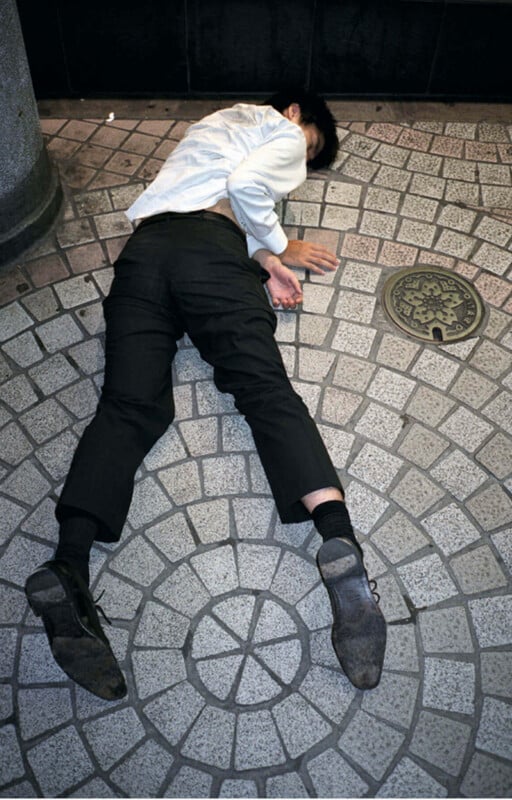 A person wearing black pants, black socks, black shoes, and a white shirt is lying face down on a tiled sidewalk, one arm extended and face turned away, near a metal utility cover.