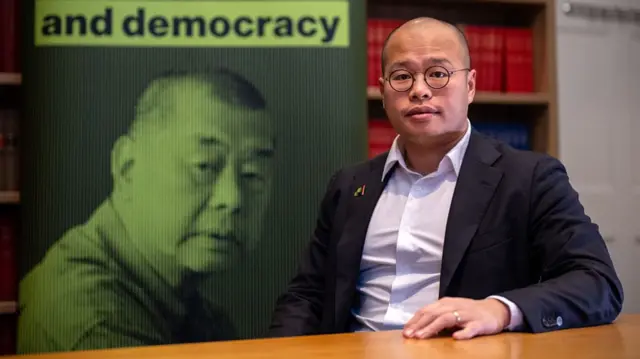 Sebastien Lai posing for a photo at a table. He's wearing a suit and behind him is a poster featuring Jimmy Lai