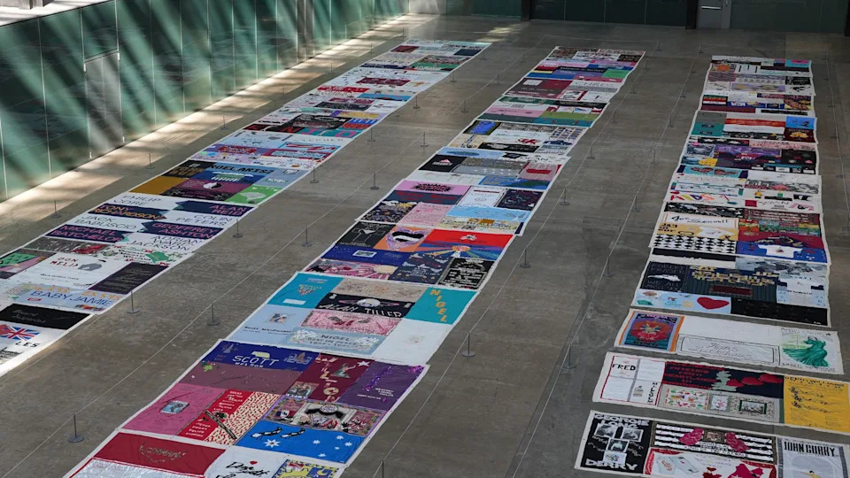 The panels and quilts of the UK Aids memorial quilt are laid out on the floor of a huge room - the Turbine Hall at the Tate Modern - in three parallel lines. They are all brightly coloured textiles with names, messages and pictures of loved ones on.