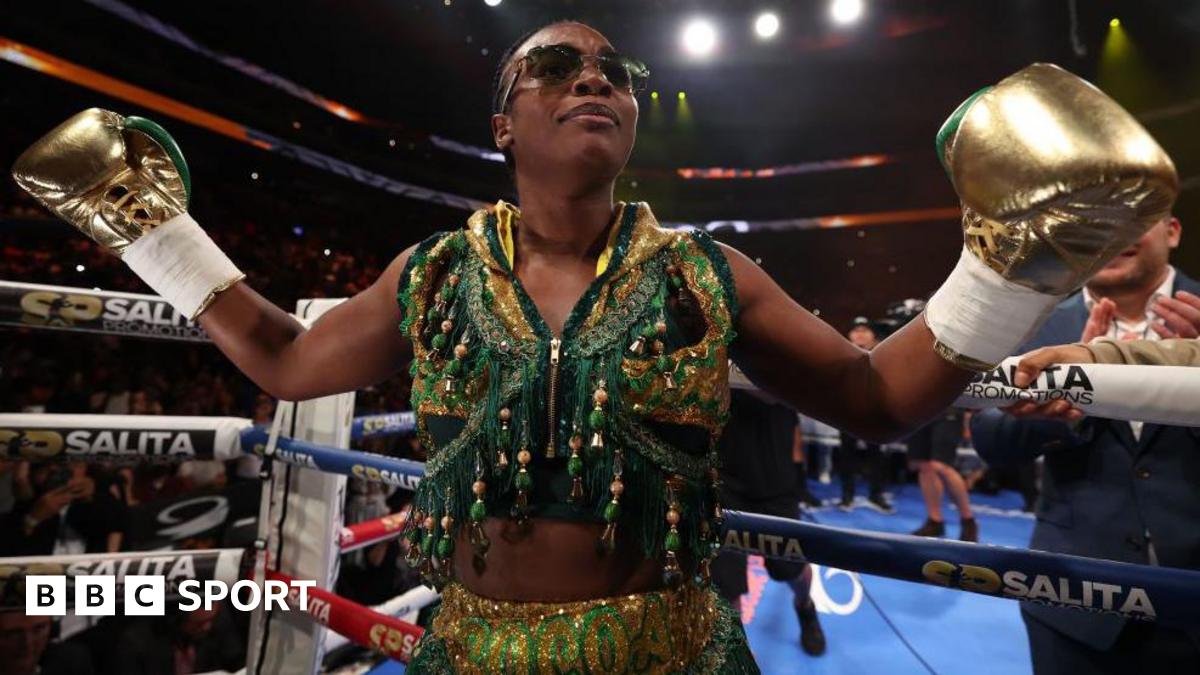 Claressa Shields holds out her arms and is wearing gold boxing gloves and a gold and green vest as she stands next to the ropes of a boxing ring