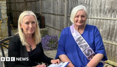Two women are sat down on chairs in a garden. They are both smiling at the camera. One women has blonde hair and it wearing a black top and jacket. She is holding a clipboard with a piece of paper that has a large heading that reads 'Waspi'. The other women has short grey hair and is wearing a blue top. She is wearing a pink sash that says 'Waspi' on it.