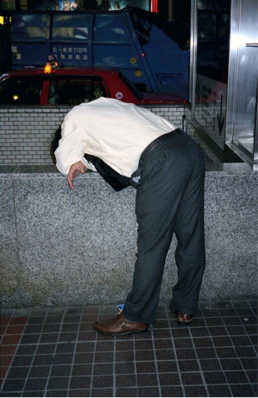 A person in dress pants and a white shirt leans over a stone wall on a city sidewalk at night, appearing exhausted or unwell. A red taxi and blue vehicle are visible in the background.