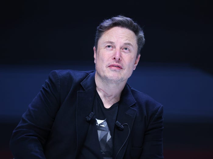 Elon Musk is in a black suit jacket and a black graphic t-shirt on stage. He is looking to the top left corner of the image.