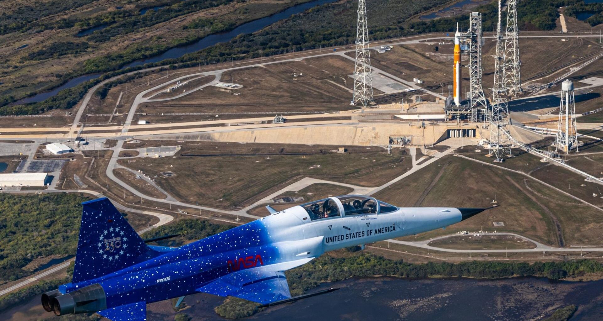 A blue and white jet flies over a launch complex where an orange and white rocket sits on a launch pad