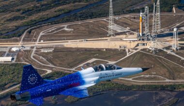 A blue and white jet flies over a launch complex where an orange and white rocket sits on a launch pad