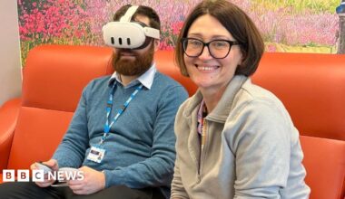 A bearded man in a blue jumper on a orange sofa wears a virtual reality headset. In the foreground, his female colleague with black bobbed hair and glasses sits smiling.