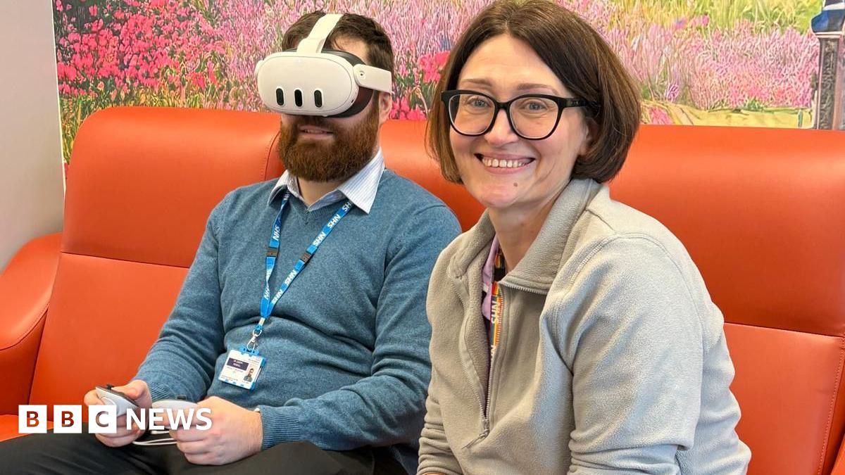 A bearded man in a blue jumper on a orange sofa wears a virtual reality headset. In the foreground, his female colleague with black bobbed hair and glasses sits smiling.