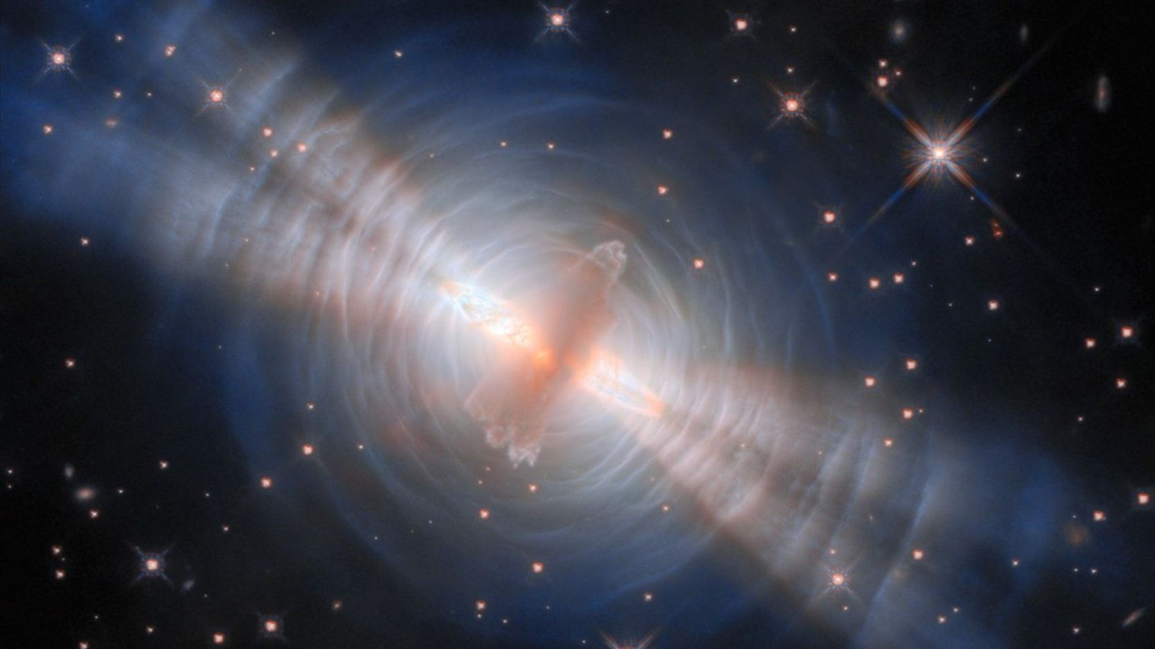 A cropped version of the Egg Nebula image from the Hubble Space Telescope. A beautiful image of space with sparkling stars in the background. There's a bright hazy structure in the center surrounded by a shell of blue-ish dust and with four streaks of light coming out of it.