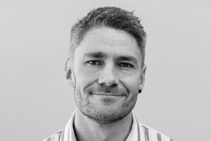 A black and white photo of a man with a stripey shirt stands smiling.