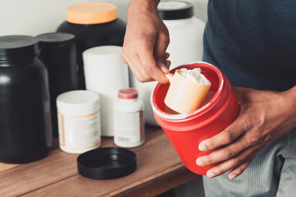 Person scooping protein powder from a red container, with various supplement bottles on a wooden table