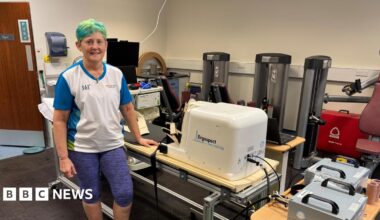 Julie Smith, with died green hair, wearing a white and blue tee-shirt and purple/black pattered leggings to just below the knee stands to camera left with her hand resting on a horizontal stepping machine. Fitness and monitoring instruments of different kind are visible around the room