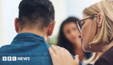 Stock image of a girl with long hair tied in a ponytail stands next to a window and looks outside, wearing a vest and checked pyjama bottoms.