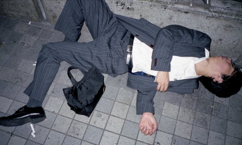 A man in a pinstripe suit lies on a tiled sidewalk, eyes closed, with a black bag and a cigarette nearby. His shirt is untucked and he appears disheveled, resting against a concrete wall.