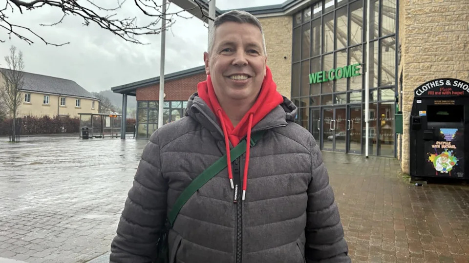 A middle aged man wearing a grey coat with a red hoodie underneath. He's smiling, and stood in front of a primary school. It says 'welcome' above the entrance to the school. Two houses are in the background. The sky is grey. 