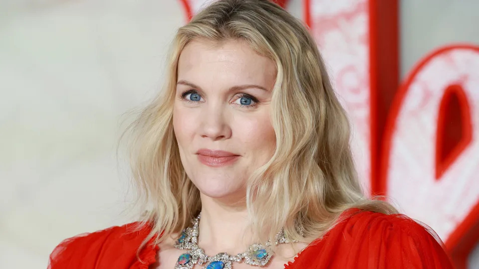 Close up of Emerald Fennell on the red carpet. She has blonde, shoulder-length hair and wears a red dress and large necklace featuring blue jewels. 