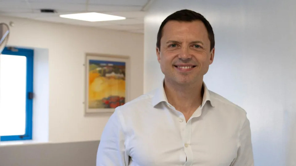 Dr Lavinio smiles at the camera while standing in a corridor. He wears a white open necked shirt and is clean shaven with short dark hair. There is a colourful painting on the wall in the distance.