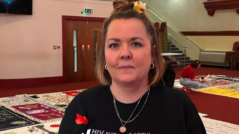Jessica Harding in a black t-shirt, with a red ribbon, standing in front of the quilt laid on the a claret-coloured floor