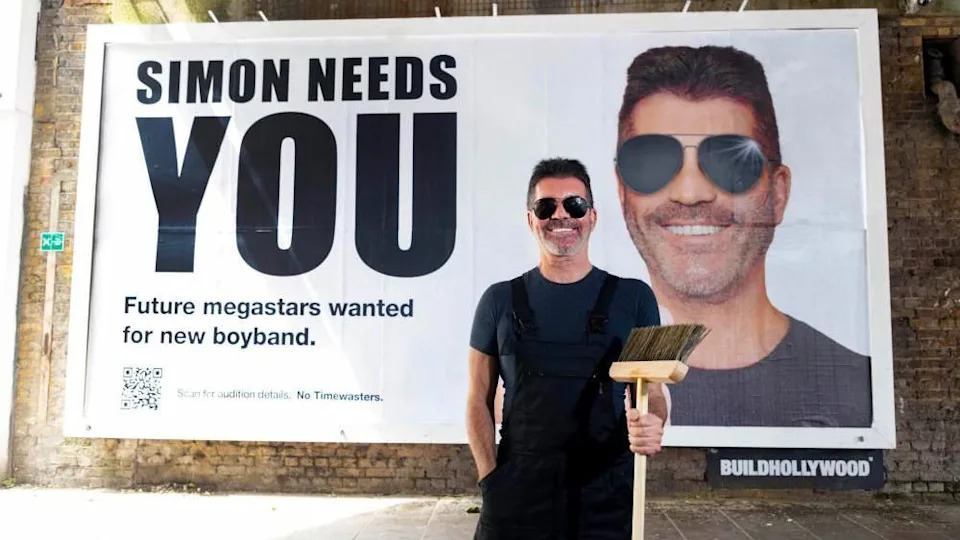 Simon Cowell is wearing sunglasses, black dungarees and a blue T-shirt and is smiling while holding a wooden broom. Behind him is a billboard with a photo of him smiling and 'Simon needs you. Future megastars wanted for new boyband'.