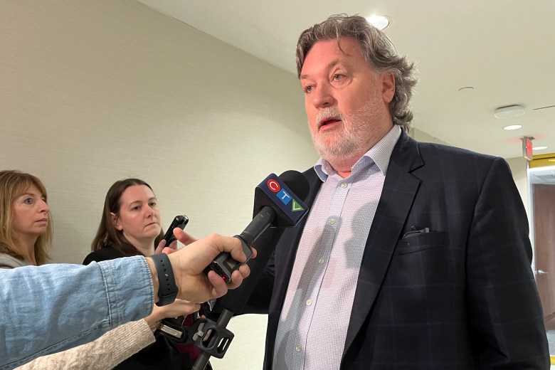 A middle aged white man with grey hair and a white beard talks to reporters.