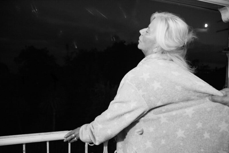 An older woman with long, white hair stands on a balcony at night, wearing a star-patterned robe. She looks up thoughtfully at the sky, with trees and a bright moon visible in the background.