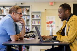 Two people sitting across from each other at a table in a library, one using a laptop while they talk. Bookshelves are visible in the background.
