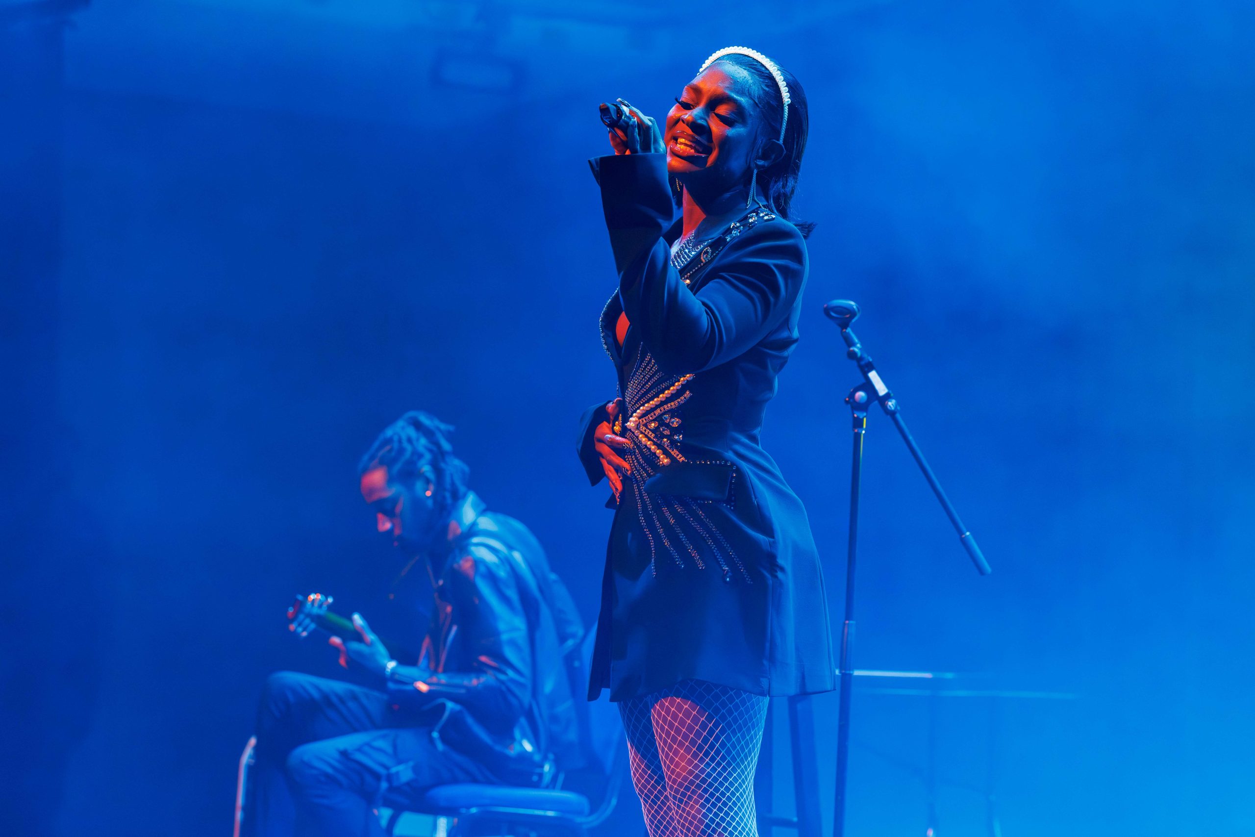 A vocalist performs onstage under blue lighting, holding a microphone while a guitarist sits nearby playing. A microphone stand and other stage equipment are visible in the background.