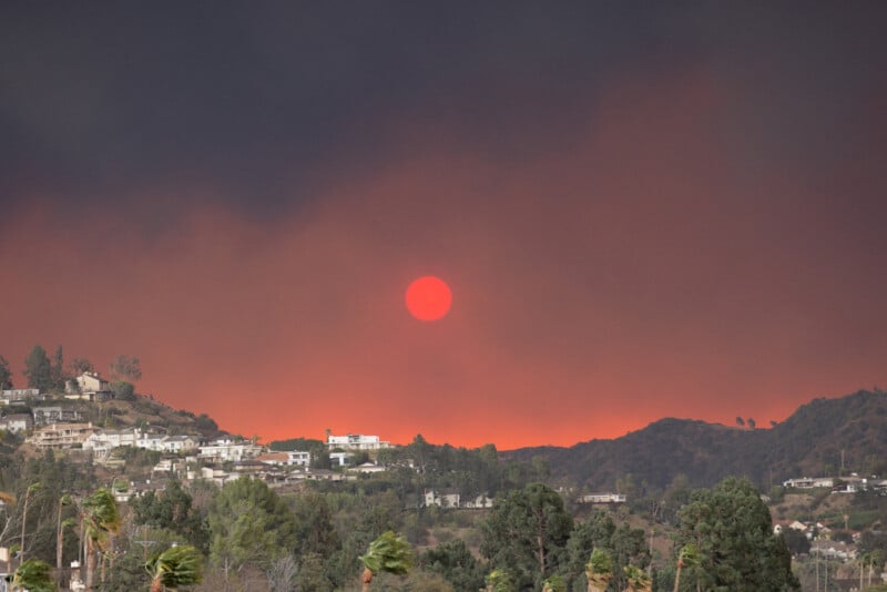 A red sun hangs in a smoky sky over hills with houses, as thick smoke and an orange glow from a wildfire cover the landscape, creating an eerie and dramatic scene.