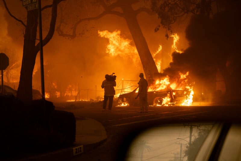 A cameraman films a firefighter standing near burning cars and large flames during a wildfire at night, with an orange glow lighting up smoke and trees; a street sign and a car mirror are visible in the foreground.